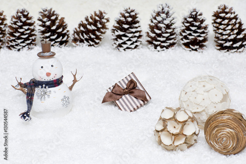 Gift box and snowman are in the snow. Christmas light background has cones. 