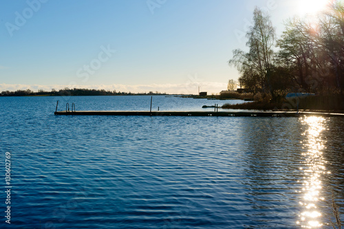 Backlit swimming pier in calm water. Sun and reflections visible over some trees. © imfotograf