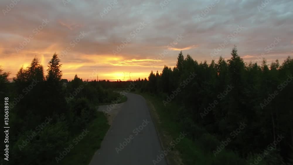 On the road in the forest, car rides with the lights on. The camera flies over the road towards the car, between the tops of the trees. Dawn and the power line on the background of beautiful sky. Aeri On the road in the forest, car rides with the lights on. The camera flies over the road towards the car, between the tops of the trees. Dawn and the power line on the background of beautiful sky. Aeri