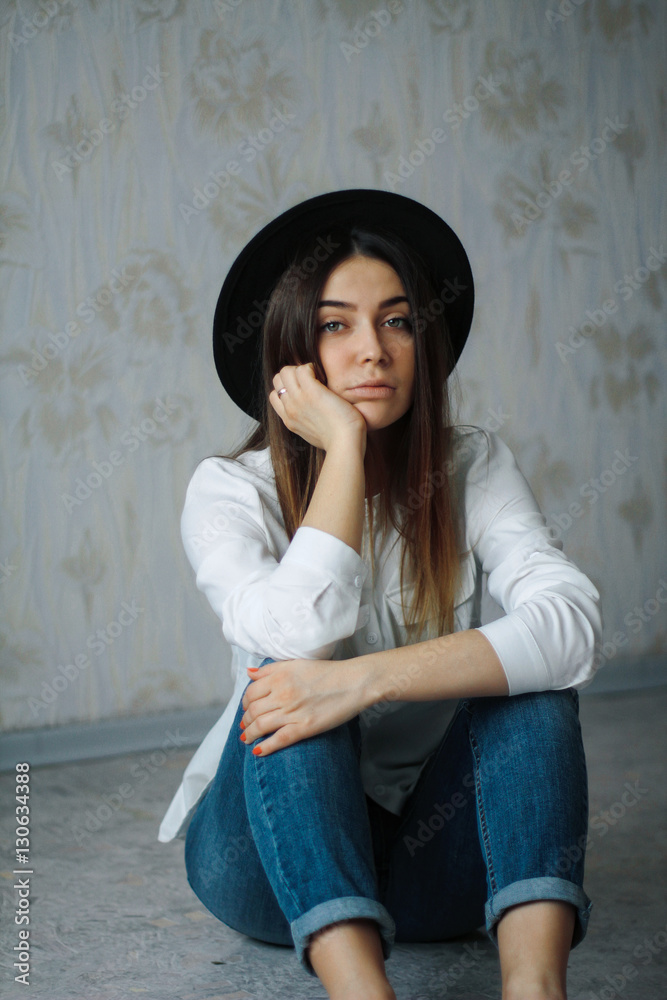 beautiful girl in a white shirt, blue jeans and a black hat sitting on the floor and looking into the camera. look sad brunette, lips inflated