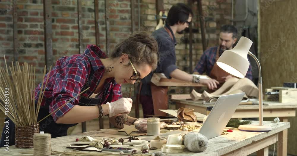 We see a busy workshop. The hipster girl on the foreground is painting wooden toys, and two men, young and middle-aged are making the cabriole table leg/Busy Hipster Workshop We see a busy workshop. The hipster girl on the foreground is painting wooden toys, and two men, young and middle-aged are making the cabriole table leg/Busy Hipster Workshop