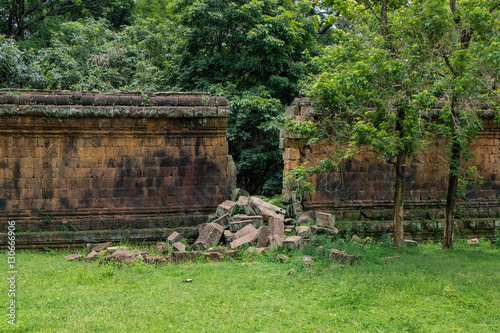 Ancient stone wall with gap from a break through, some loose stone blocks from the breach are visible in front. The wall is located in the antique ruins of Angkor.