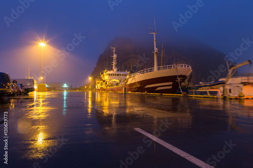 Ship docked in foggy harbor on the Westman islands, Iceland. Night scene with light reflection from streetlamps on wet pavement in misty weather © Sander