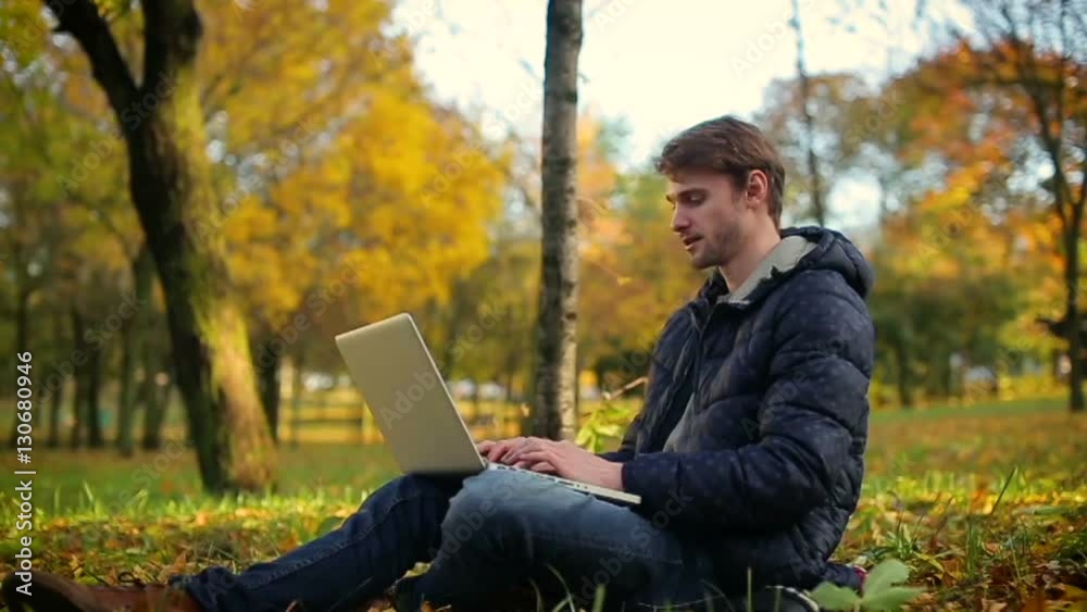 man using laptop in autumn park working sitting on the grass, dolly camera/it startup programmer man working in the park using laptop sitting on the grass in sunny autumn day slowmotion slider cam man using laptop in autumn park working sitting on the grass, dolly camera/it startup programmer man working in the park using laptop sitting on the grass in sunny autumn day slowmotion slider cam