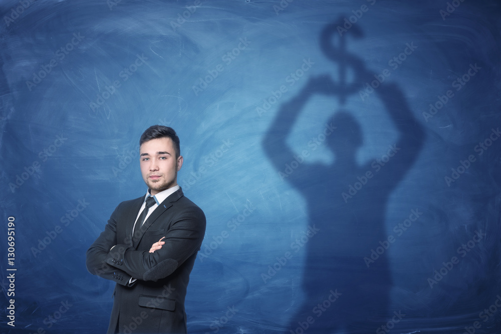 Businessman standing and shadow on the blackboard behind him holding dollar sign above his head Businessman standing and shadow on the blackboard behind him holding dollar sign above his head