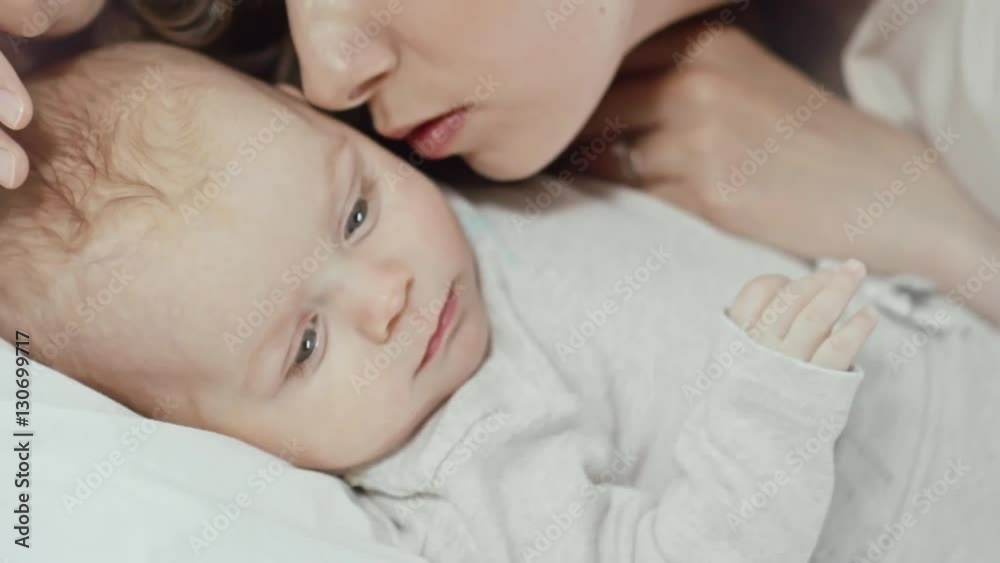 Handheld shot of young beautiful mother gently kissing her baby son lying on bed Handheld shot of young beautiful mother gently kissing her baby son lying on bed