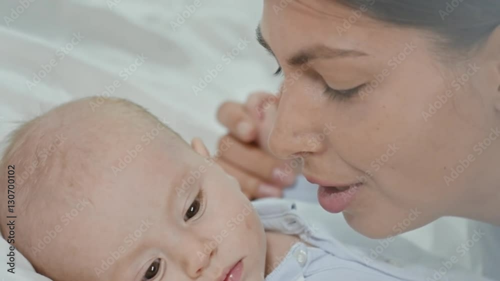Mother talking to little baby boy lying on bed, then rubbing off lipstick stains of his cheek Mother talking to little baby boy lying on bed, then rubbing off lipstick stains of his cheek