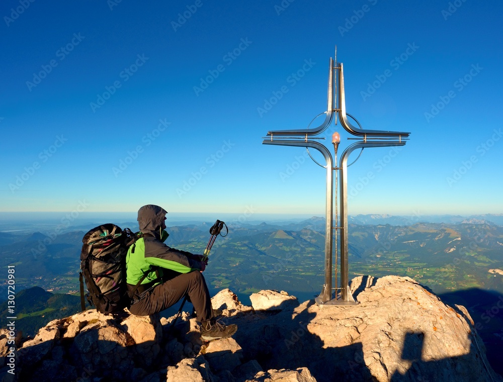 Tired hiker sit bellow crucifix on mountain peak. Iron cross at Alps mountain top.