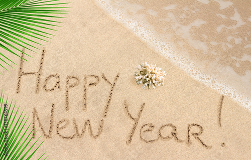 Happy New Year handwritten on sandy beach with ocean wave and palm leaves on background
