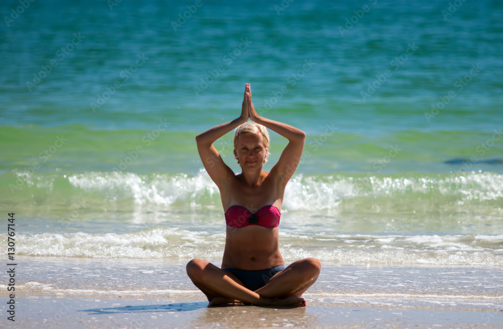 Middle-aged woman practicing yoga at the beach against the backdrop of blue sea. Selective focus.
