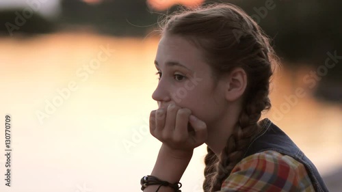 Sad teen girl (14-15) resting chin in hand thinking outdoor. Portrait of pensive teenager looking away sitting by calm lake at sunset.