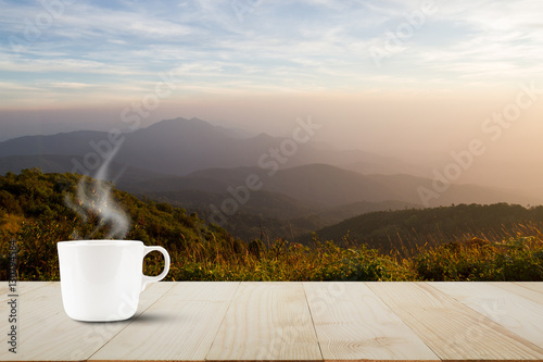 Hot coffee cup with steam on vintage wooden table top on blurred meadow and foggy mountain background during sunrise