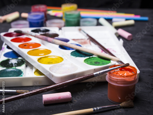 Bright multi-colored paint, pastels, crayons, pencils, brushes on a black wooden background. Set for the artist. Education.