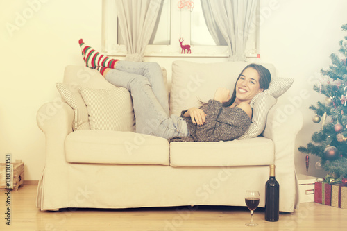 Beautiful woman lying on sofa next to Christmas tree enjoying glass of wine
