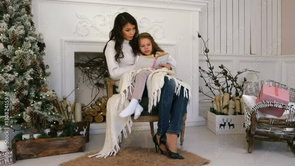 Young mother and her little daughter reading a Christmas tale relaxing in an armchair by the fire place