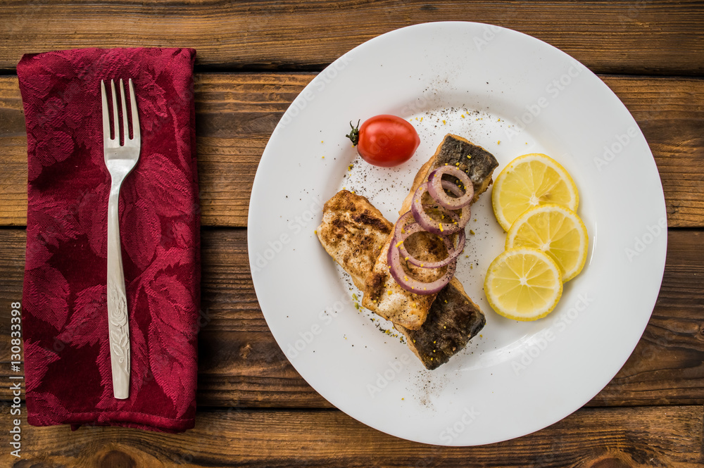 Fried fish fillet with lemon and tomatoes on a white plate. Wooden background. Top view. Close-up Fried fish fillet with lemon and tomatoes on a white plate. Wooden background. Top view. Close-up