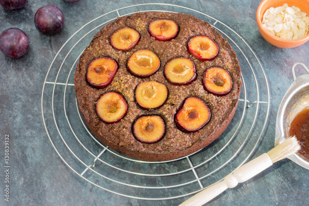 Freshly baked sticky chocolate plum cake on cooling rack surrounded by whole plums, almond slivers in a bowl and heated apricot jam in a bain-marie with pasty brush on the edge. 