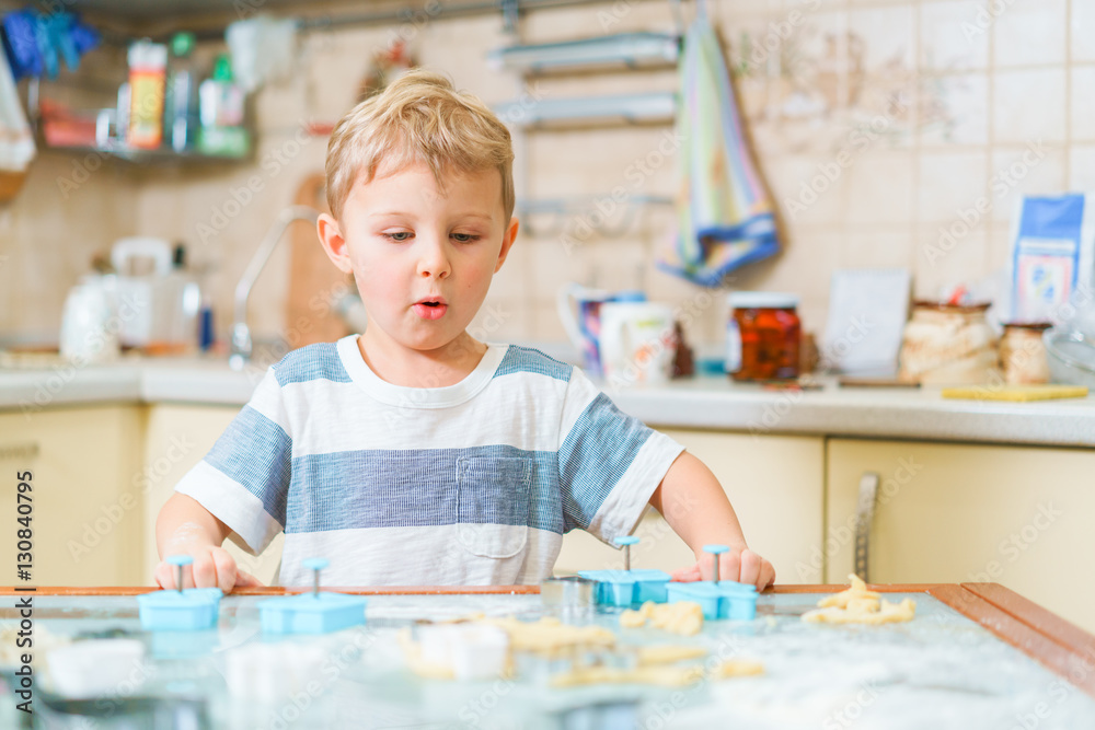 Little blond kid plays with molds for making ginger biscuits or cookies, sitting at the kitchen table with raw dough and wheat flour. Rapt face expression. Little blond kid plays with molds for making ginger biscuits or cookies, sitting at the kitchen table with raw dough and wheat flour. Rapt face expression.