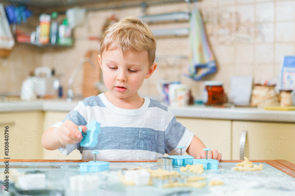Fototapeta premium Little blond kid plays with molds for making ginger biscuits or cookies, sitting at the kitchen table with raw dough and wheat flour. Rapt face expression.