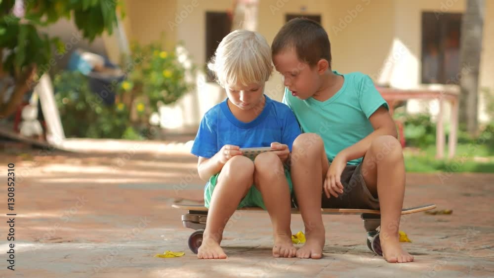 Multiracial сhildren playing online games on smart phone sitting on skateboard on backyard in summer holidays. Enjoying of using a portable handheld device in childhood. Chatting on mobile. Pan shot
