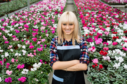 Cheerful charming woman gardener standing against large flower plantations, looking at camera.