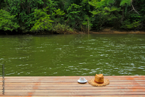 Take a relaxing time with a cup of coffee and straw hat with copy space on vacation day or weekend. Hot tea drinking on brown wooden floor with river view and tree background. Fresh air among nature. photo