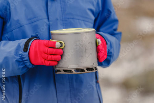 Gloved hands hold modern lightweight titanium pot for cooking on the stove and a fire during hike. Equipment for trekking