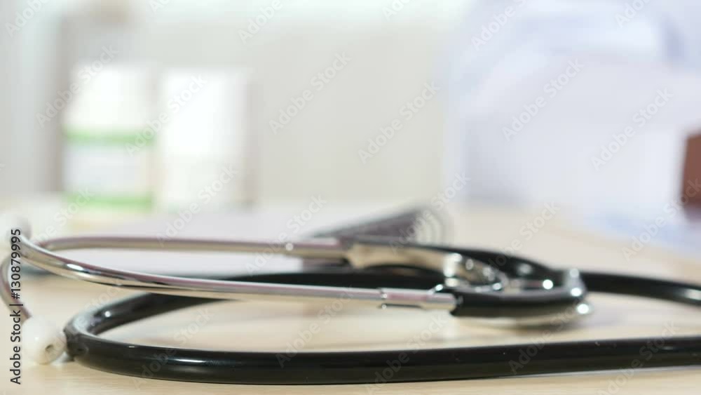 Stethoscope on table, jar of blue capsules near laptop, hands of male in white coat writing on blurred background, working as doctor at desk, prescriptions. Close up, shallow DOF, 4K Ultra HD. Stethoscope on table, jar of blue capsules near laptop, hands of male in white coat writing on blurred background, working as doctor at desk, prescriptions. Close up, shallow DOF, 4K Ultra HD.