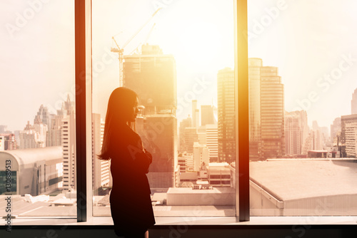 Silhouette of success businesswoman by office window with cityscape in background with sunlight. © interstid