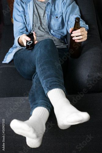 Having rest and drink on couch. Person in white socks and casual denim wear relaxing on sofa with a bottle of beer and holding TV remote control