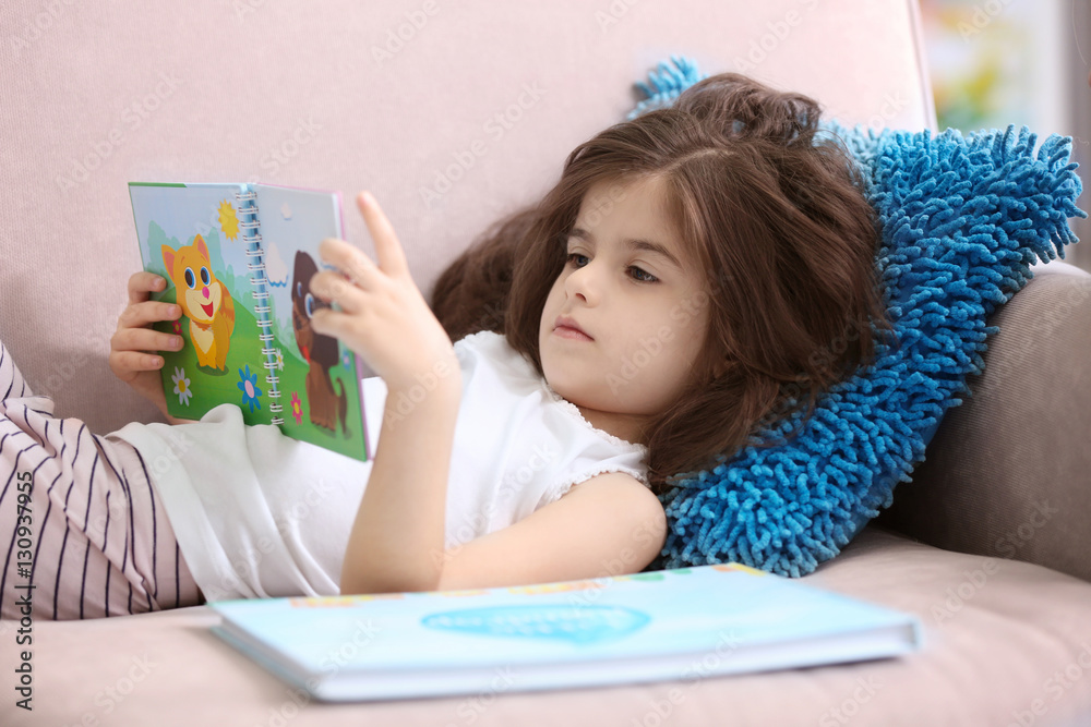 Funny little girl reading book while lying on sofa at home Funny little girl reading book while lying on sofa at home
