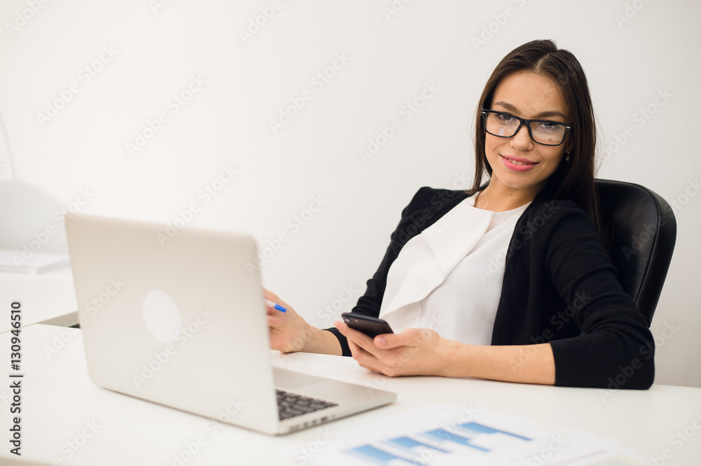 Good business talk. Cheerful young beautiful woman in glasses talking on mobile phone and using laptop with smile while sitting at her working place Good business talk. Cheerful young beautiful woman in glasses talking on mobile phone and using laptop with smile while sitting at her working place