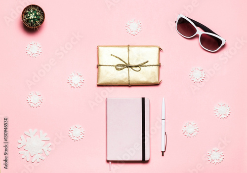 Flat lay, top view Christmas winter decorated table. Feminine desk workspace with snowflakes, pink notebook, white handle, white glasses, gift box on pink background.