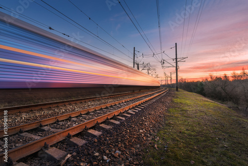 High speed passenger train in motion on railroad track at sunset. Blurred commuter train. Railway station against colorful sky. Railroad travel, railway tourism. Industrial landscape in the evening