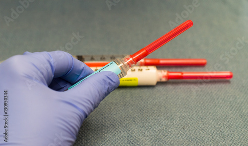 Φωτογραφία Left hand holding a syringe of pain medicine with two other syringes with red capped needles containing anesthesia drugs
in the background