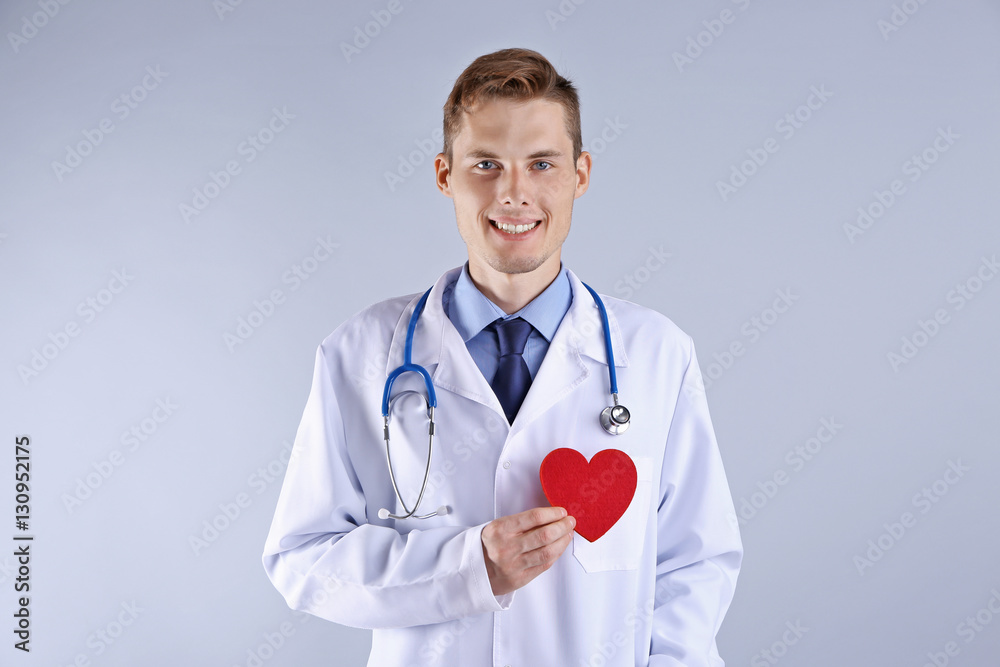 Handsome young doctor holding red heart on light background Handsome young doctor holding red heart on light background