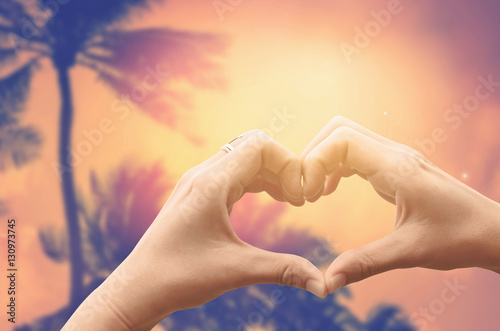 Female hands heart shape on blur tropical palm tree with sun light abstract background.
