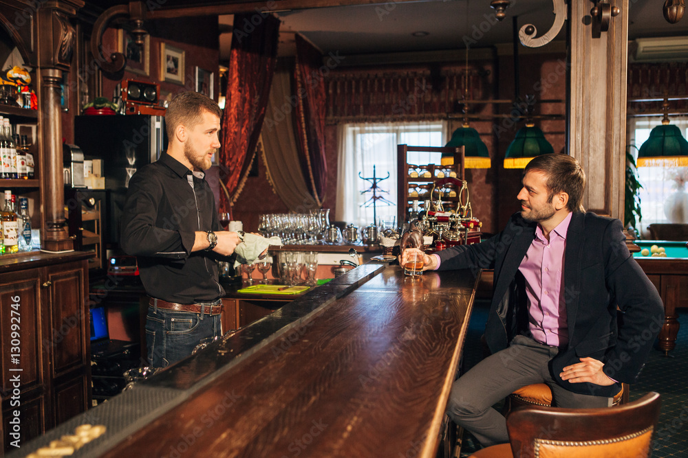 Smiling attractive young barman wiping glasses and talking to man in bar