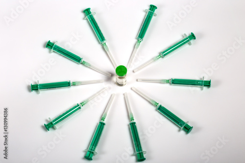 Green medical syringes are arranged on a circle. Ampule with vaccine in the center of the circle. White background, shadows 