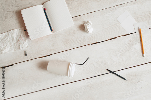 Workplace of untidy students flat lay, free space. Top view on blank copybook, pencils, coffee-to-go cup and paper wads scattered on white wooden table. Mess, disorder, bad form concept