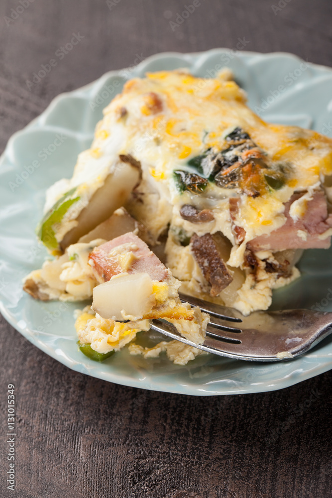 Egg breakfast casserole with potatoes, ham, mushrooms and cheese with fork on a dark wooden background