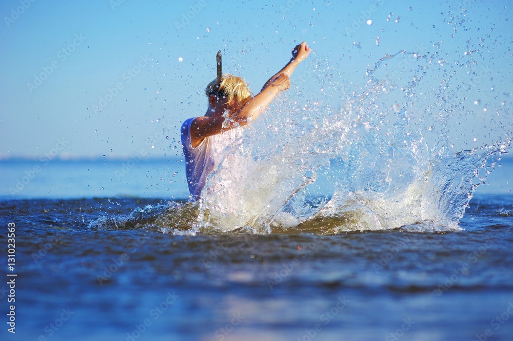 Young cheerful blonde girl enjoys swimming in the sea. She splashing in water and transparent spray scatter in different directions, creating a beautiful background.