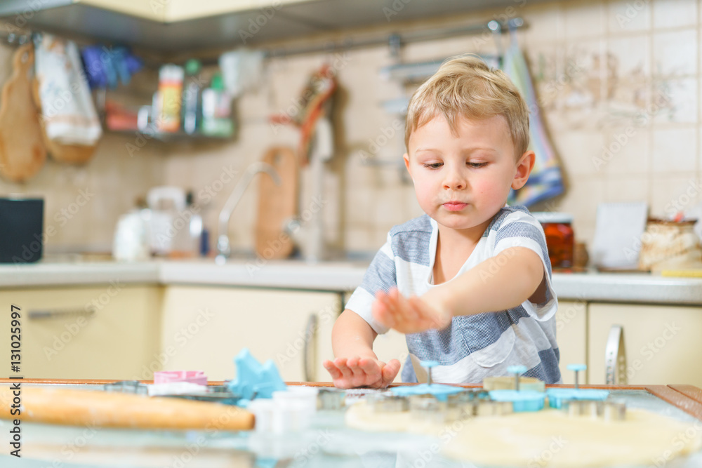 Fototapeta premium Little blond kid plays with molds for making ginger biscuits or cookies, sitting at the kitchen table with raw dough and wheat flour. Rapt face expression. Looking ahead.