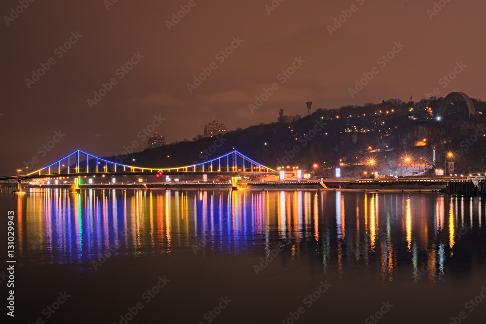 Beautiful illumination of Pedestrian Bridge reflected in the water. Evening view on the promenade along the Dnieper