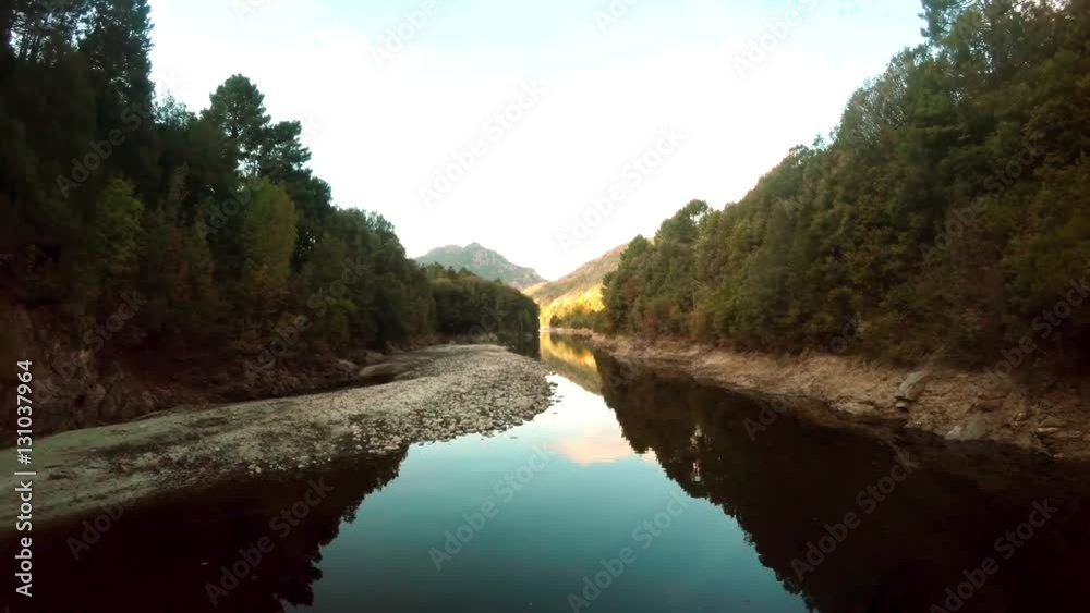 Aerial low flying bird view flying underneath small bridge in backwards flight beautiful mountain landscape river reflecting sky with rocks and trees on both sides more mountains in background 4k