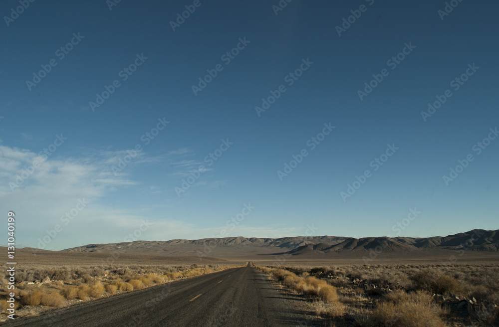 View of a straight section of the park road in Death Valley National Park, California. There is arid desert's vegetation on the side, smooth hills to close the horizon and a clear blue sky above.