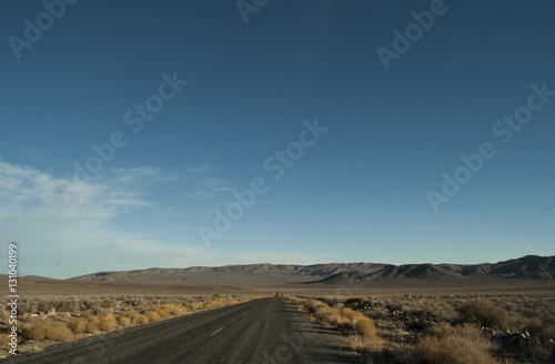 View of a straight section of the park road in Death Valley National Park  California. There is arid desert s vegetation on the side  smooth hills to close the horizon and a clear blue sky above.