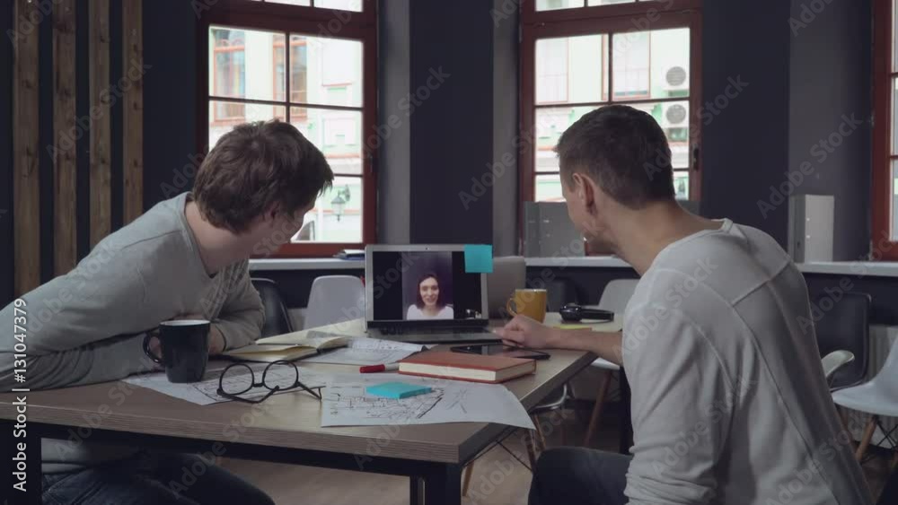 Two businessman in the modern office startup company have video conference call with colleague or friend of clent. Men using application looking on the screen laptop talking with woman. Casual