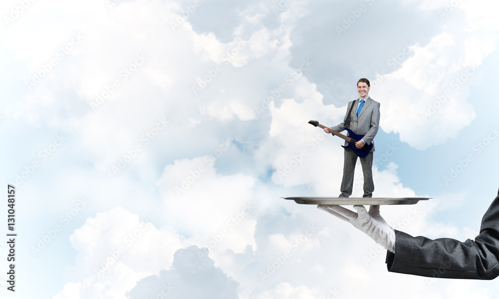 Businessman on metal tray playing electric guitar against blue sky background Businessman on metal tray playing electric guitar against blue sky background