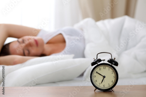 Alarm clock standing on bedside table has already rung early morning to wake up woman in bed sleeping in background. Early awakening, not getting enough sleep, oversleep, time line concept
